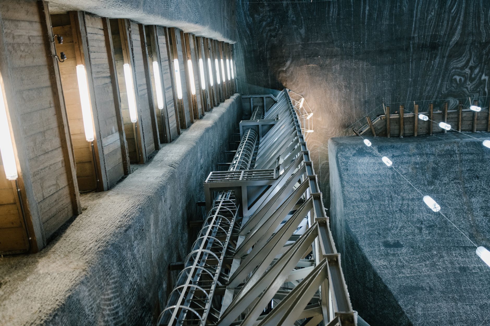 A stark, modern illustration of a deep mine shaft with heavy concrete blocks suspended by industrial cables, illuminated by harsh artificial light against a dark background.